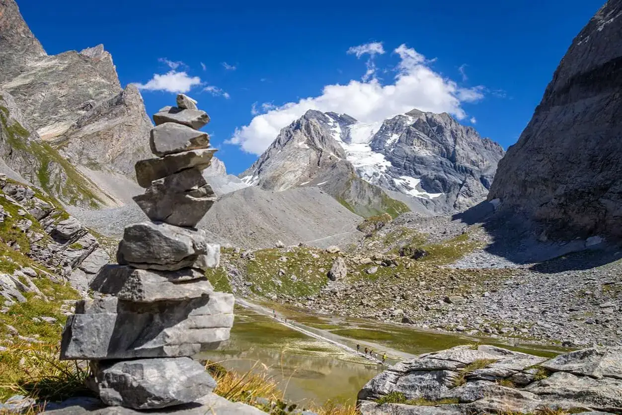 Un cairn, une pile de pierres, marque le sentier dans un paysage de montagne spectaculaire, avec un glacier au loin.