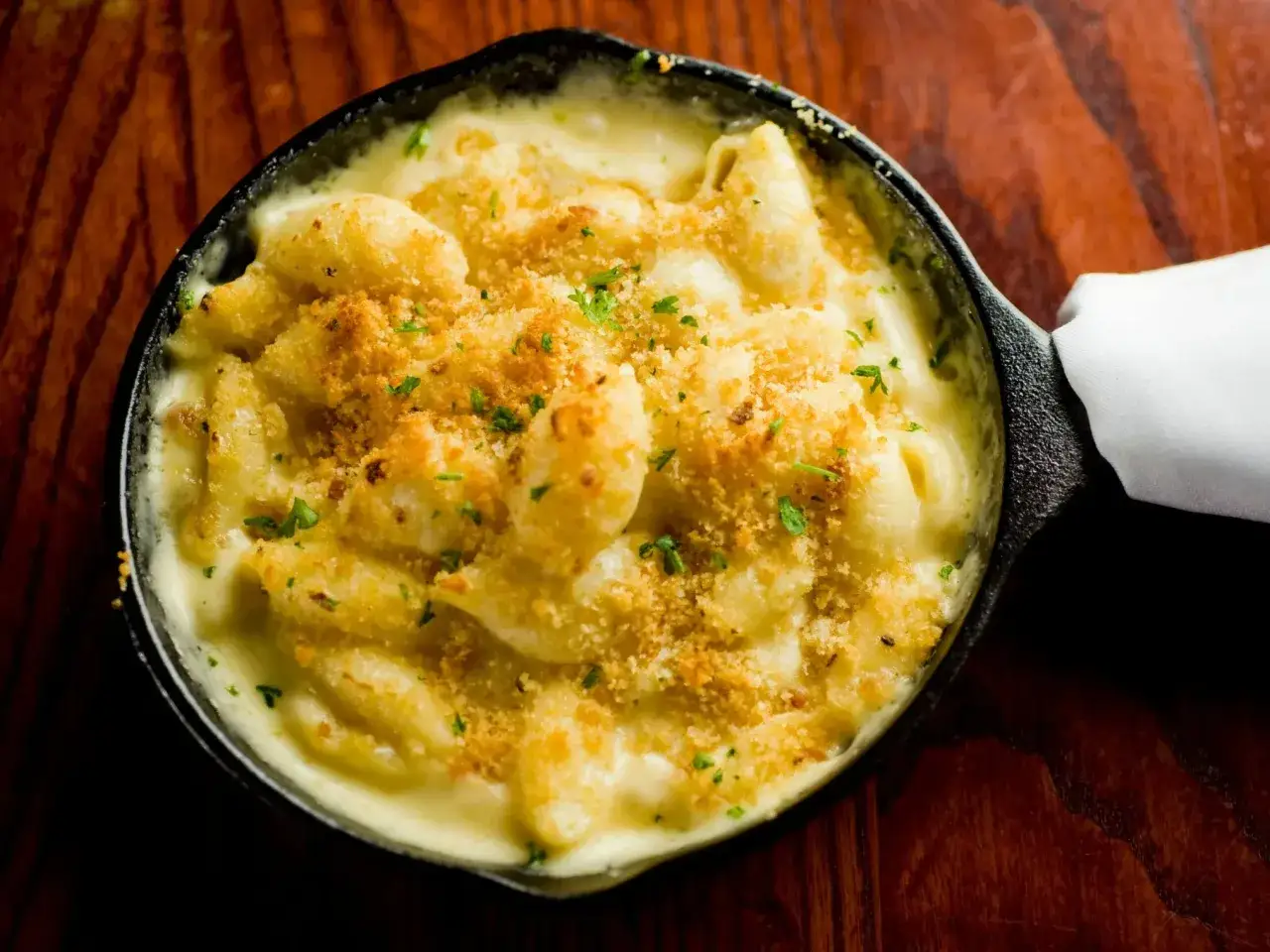 Pyszne mac n cheese w żeliwnej patelni, posypane chrupiącą panierką i natką pietruszki, na drewnianym stole.
