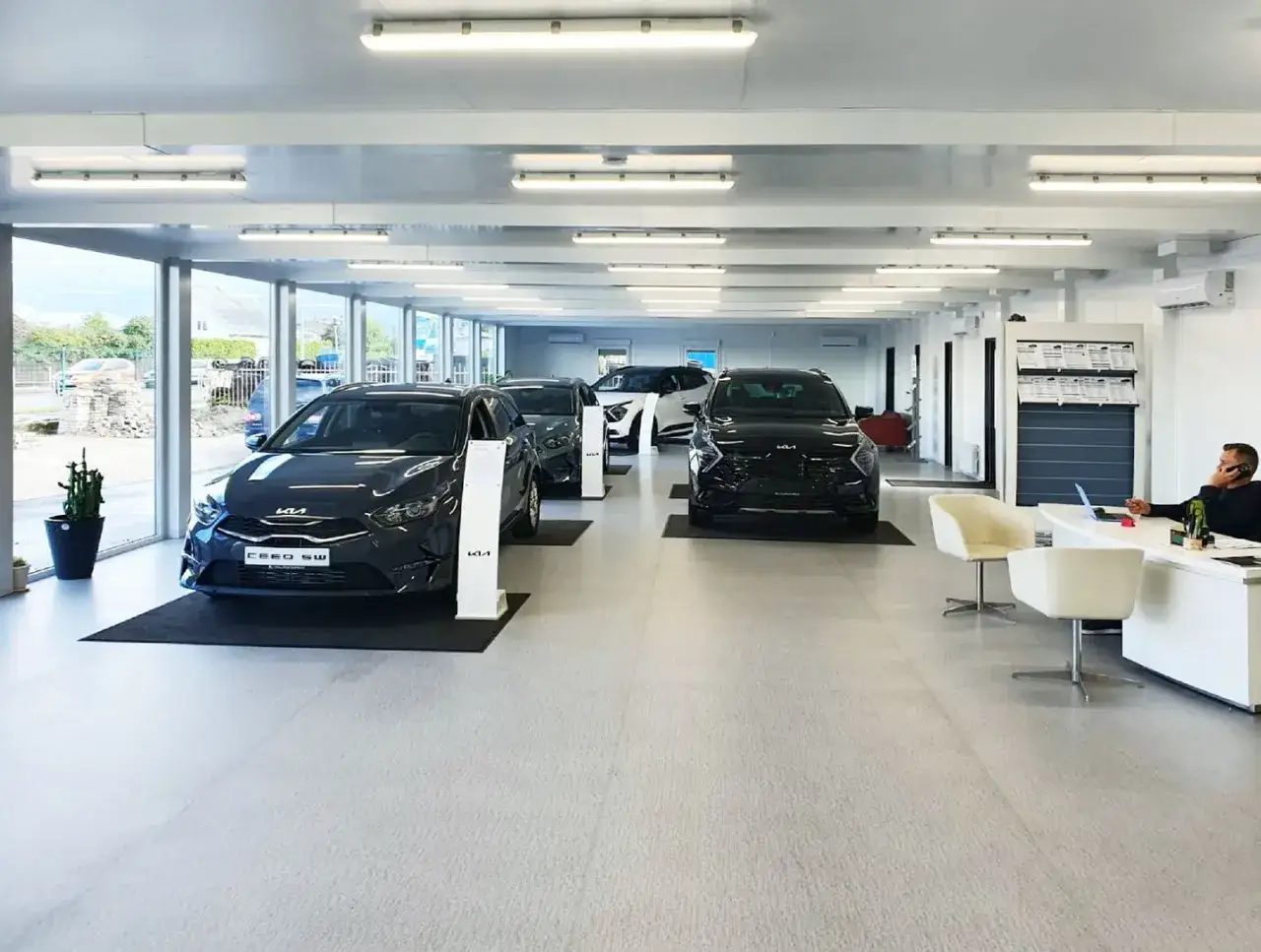 Intérieur d'un concessionnaire automobile moderne, avec plusieurs voitures neuves alignées. Un homme est au téléphone à son bureau, prêt à gérer votre **dépôt vente véhicule**.