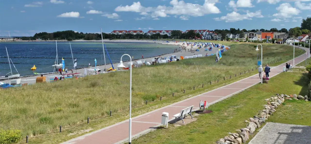 Ein sonniger Tag an der **Glowe Ostsee**. Boote liegen am Strand, Menschen entspannen sich unter Sonnenschirmen, und ein Weg lädt zum Spaziergang ein.