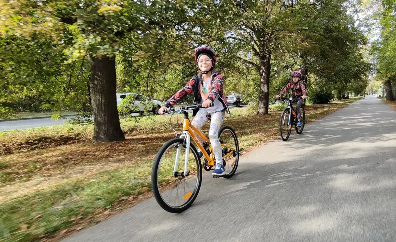 Uśmiechnięta dziewczynka na rowerze dla 7 latki, jedzie ścieżką wśród drzew.