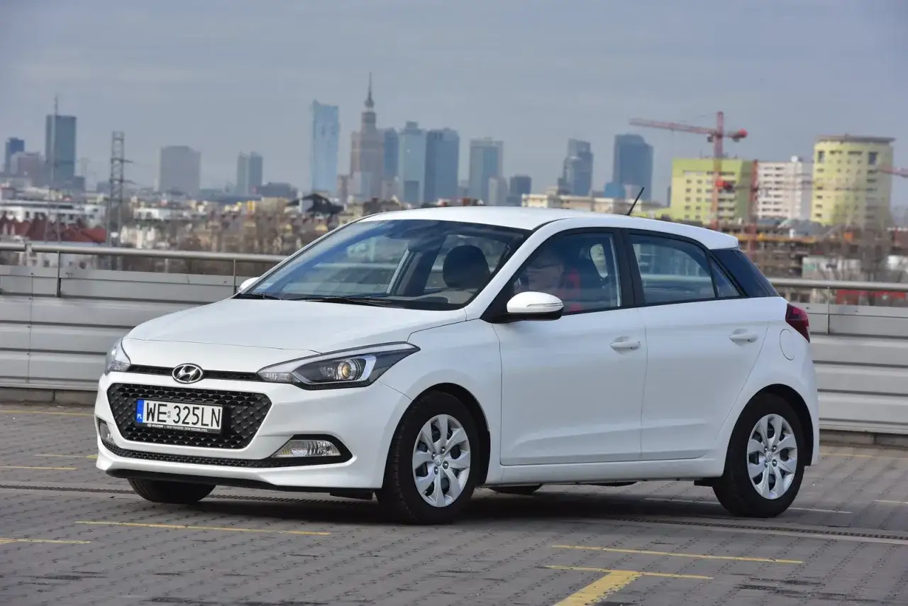 Biały Hyundai i20 z nowoczesnymi światłami, gotowy do jazdy. W tle miejski krajobraz.