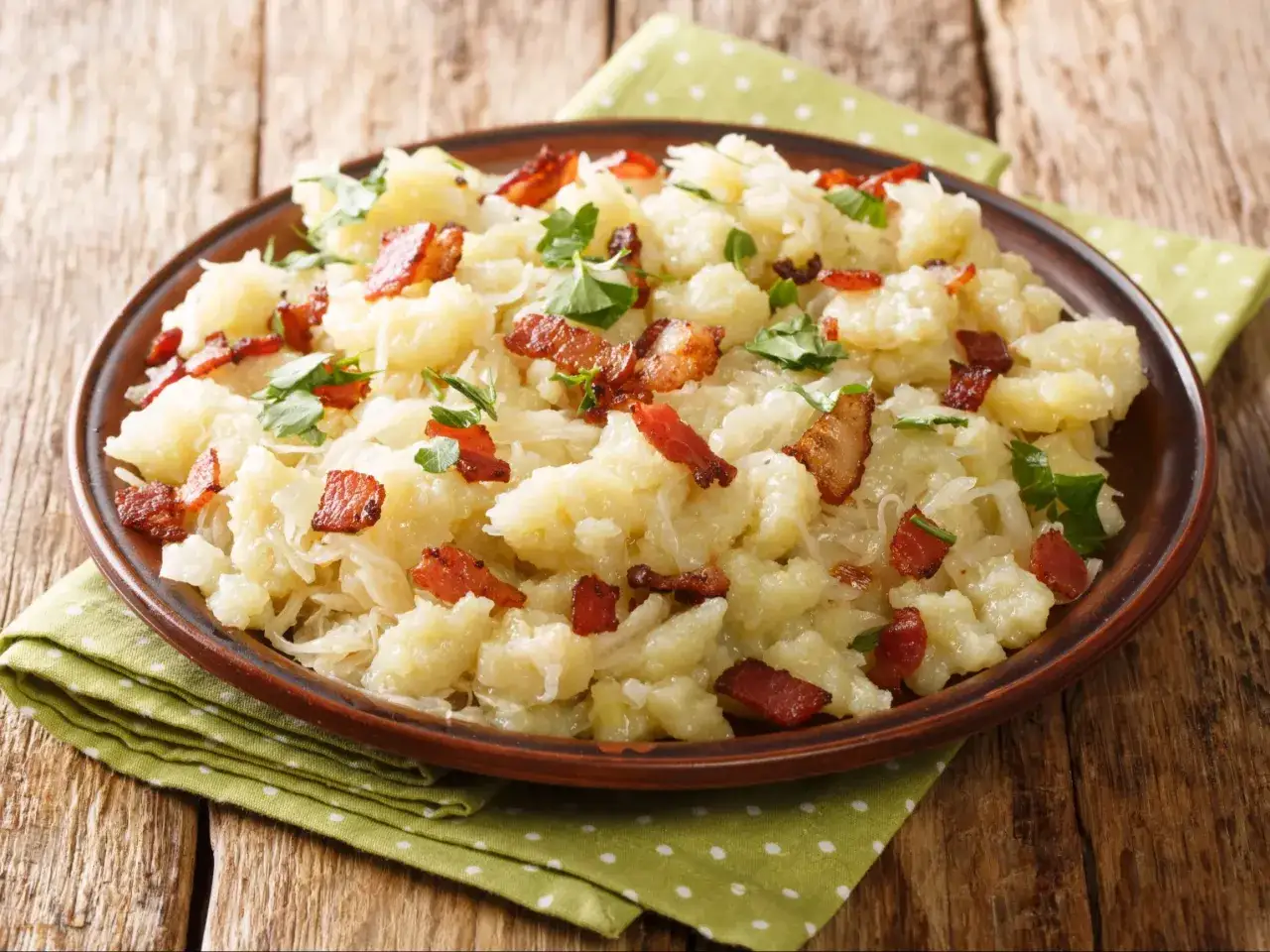 Szare kluski wielkopolskie z chrupiącym boczkiem i natką pietruszki na brązowym talerzu, podane na zielonej serwetce w kropki.