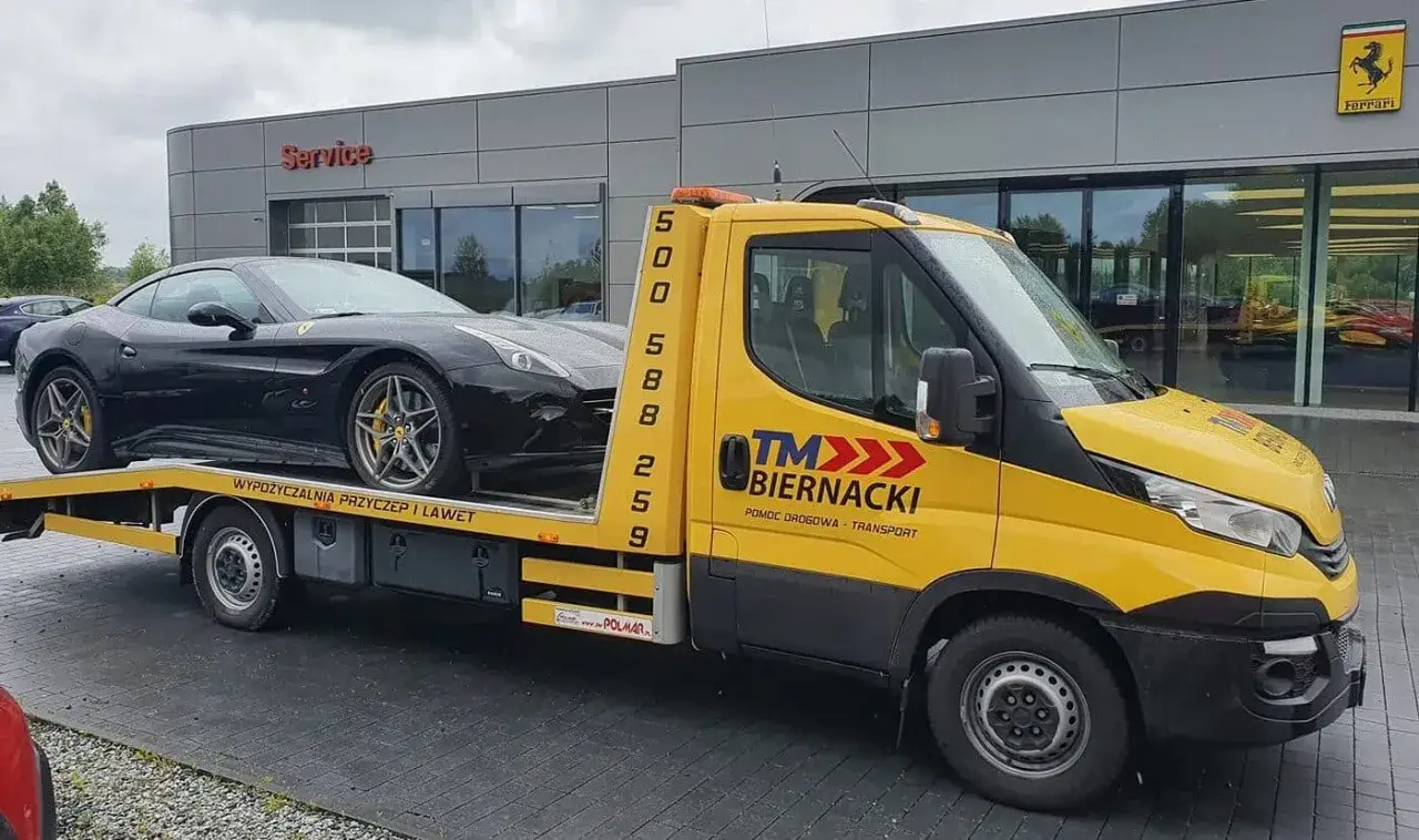 Żółty pojazd specjalny pomoc drogowa transportuje czarnego Ferrari. Przepisy dotyczące transportu luksusowych aut są surowe.