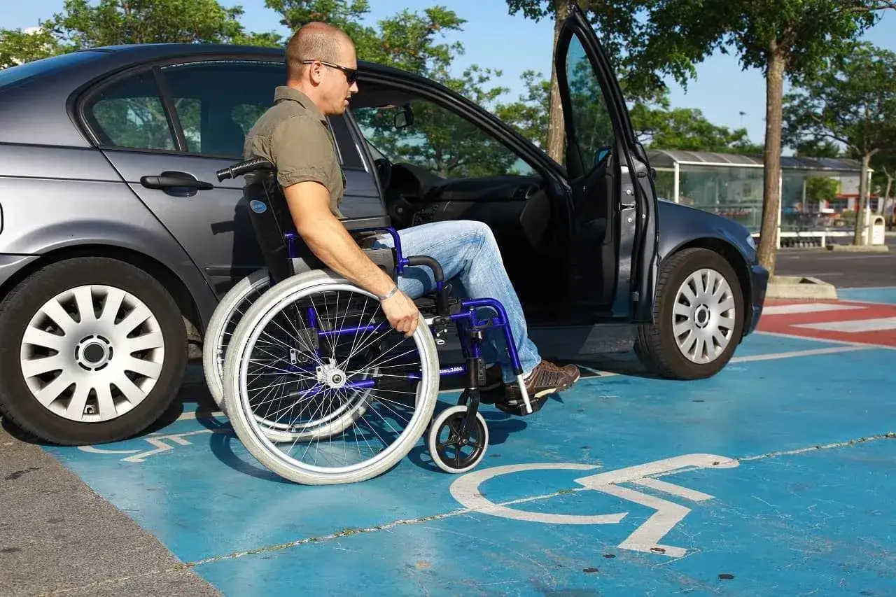 Mężczyzna na wózku inwalidzkim wsiada do samochodu na miejscu parkingowym dla niepełnosprawnych. Posiadanie prawa jazdy dla niepełnosprawnych otwiera nowe możliwości.