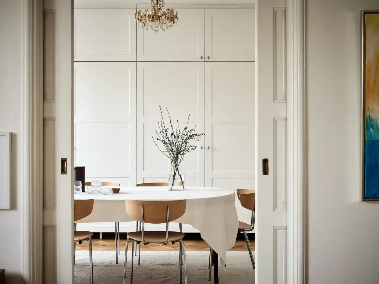 Comedor con mesa cubierta, sillas de madera y un gran armario empotrado en blanco roto NCS. Una l&aacute;mpara de ara&ntilde;a dorada ilumina la escena.