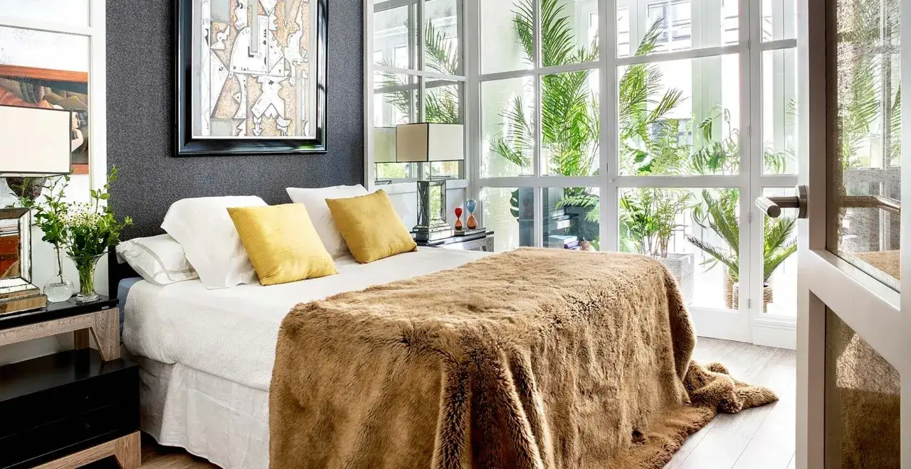 Ideas decoracion habitacion: cama acogedora con manta peluda, almohadas amarillas y arte abstracto sobre pared oscura, junto a ventana con plantas.