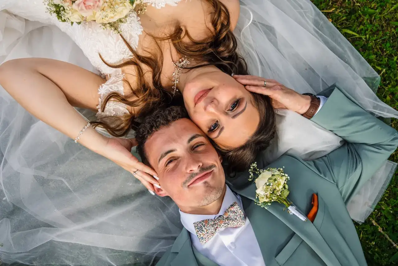 La photo de mariage capture un couple heureux, allongé sur l'herbe, le visage de la mariée près de celui du marié.