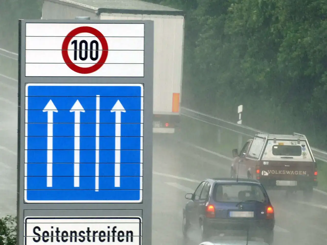 Verkehrsschilder zeigen 100 km/h und drei Fahrspuren. Autos und ein LKW fahren bei Regen, der Seitenstreifen ist befahren.