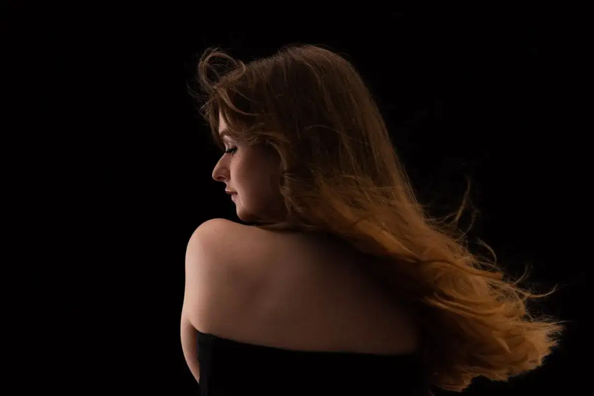 Une jeune femme aux cheveux roux longs et ondul&eacute;s, vue de profil, se d&eacute;tache sur un fond noir. L'effet de **mettre un fond noir sur une photo** accentue la douceur de sa peau et la texture de ses cheveux.