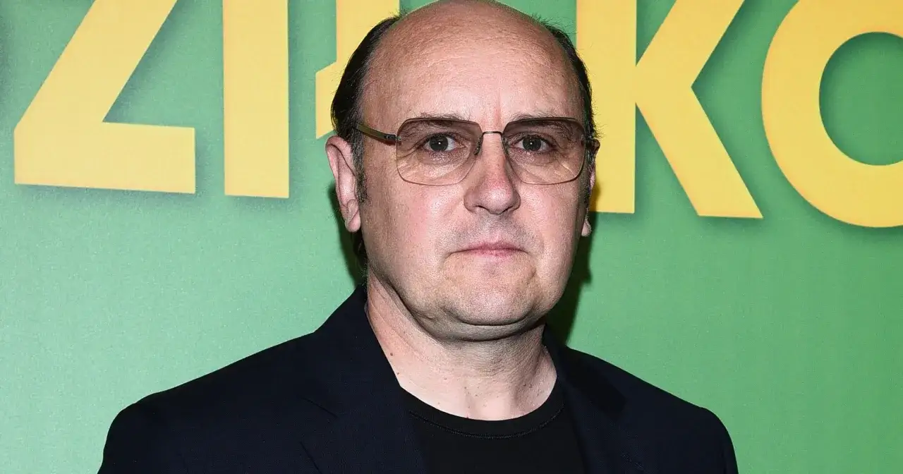 Adam Woronowicz w okularach na tle zielonej ściany z żółtymi literami. Jego charakterystyczna twarz znana z wielu polskich filmów.