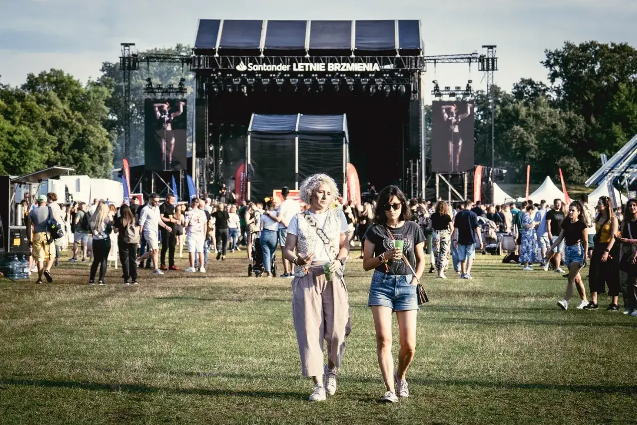 Dwie kobiety idą przez trawiasty teren festiwalu Santander Letnie Brzmienia we Wrocławiu. W tle widać scenę i tłum ludzi.