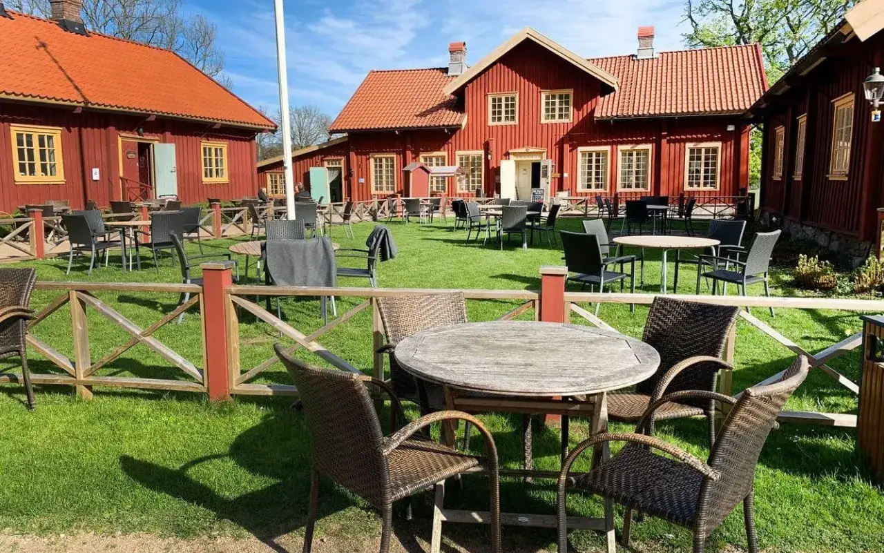 Röda trähus med vita knutar och orange tak. Uteservering med bord och stolar på gräsmattan. Perfekt för en fika och en kaffetår.