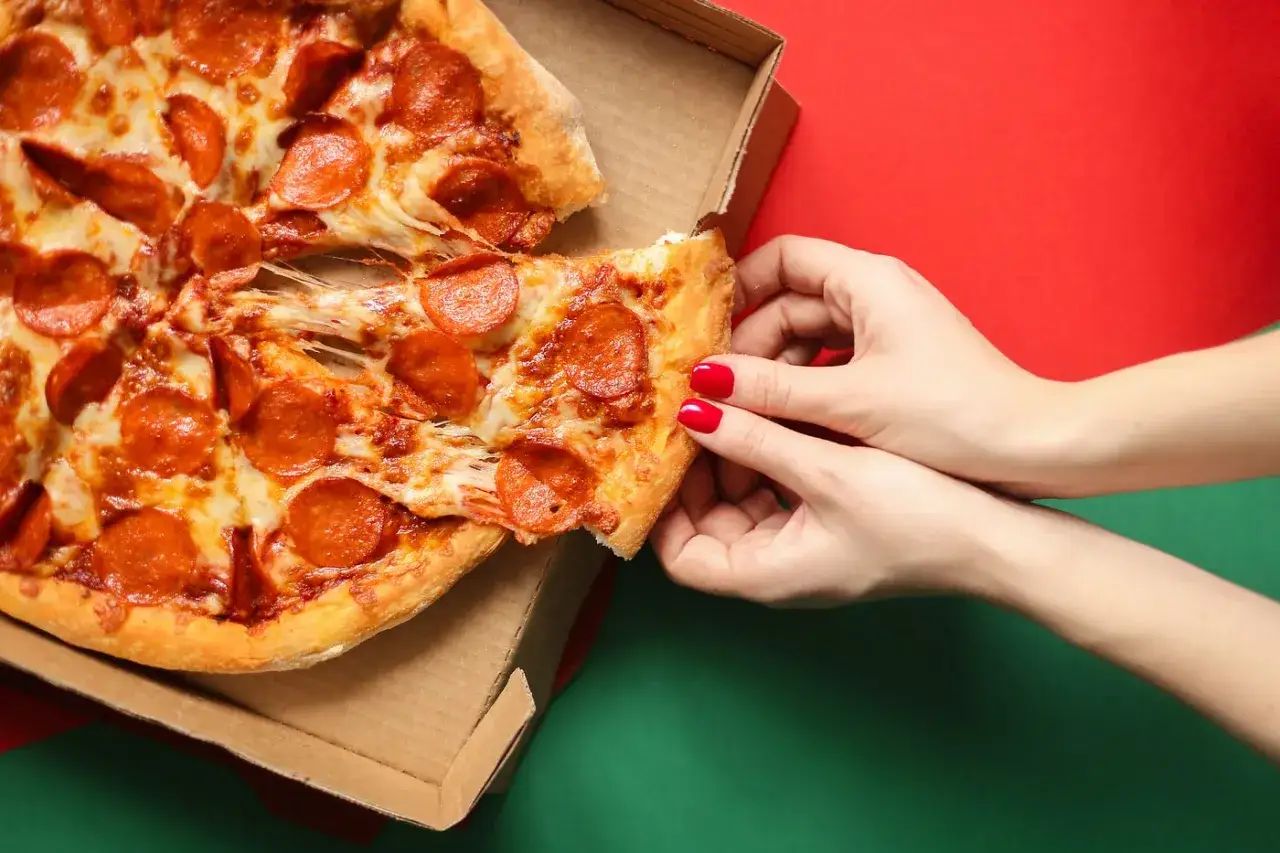 Hände greifen nach einem Stück Pepperonipizza aus einer Pappschachtel. So isst man Pizza richtig – mit viel Käse und leckeren Salamischeiben.