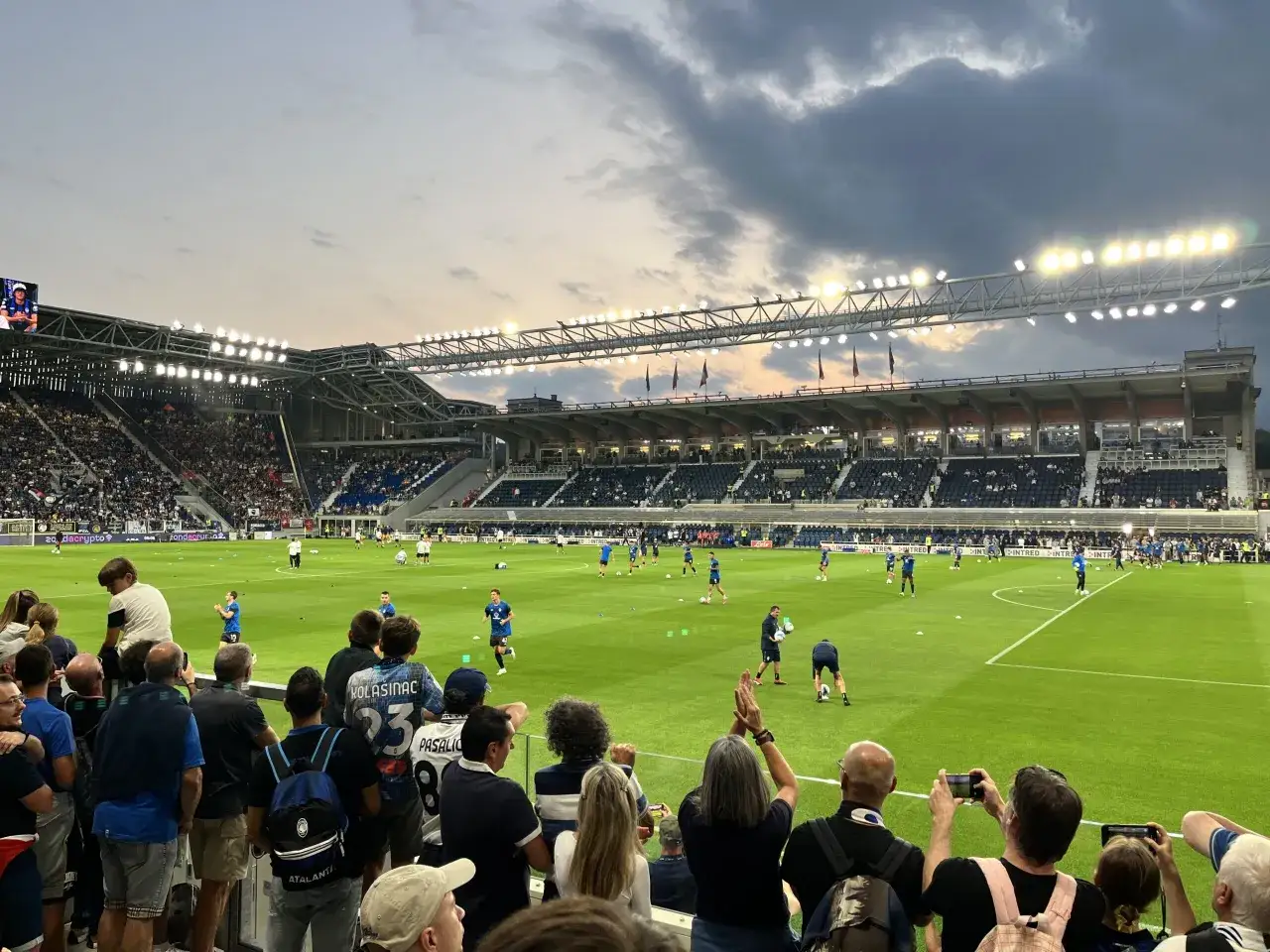 Piłkarze z Bergamo rozgrzewają się na murawie stadionu. Kibice na trybunach obserwują ich przygotowania do meczu.