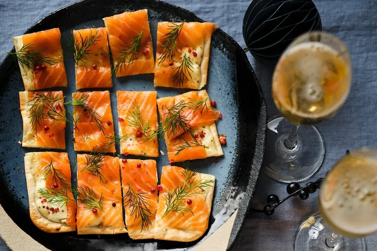 Petits flamenkuch saumon, garni d'aneth et de baies roses, servi sur une assiette bleue avec des coupes de champagne.