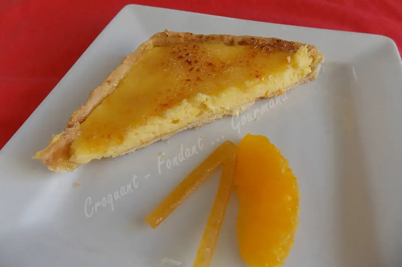 Une part de tarte à l'orange, avec sa garniture dorée et caramélisée, est servie sur une assiette blanche avec une tranche d'orange et des bâtonnets confits.