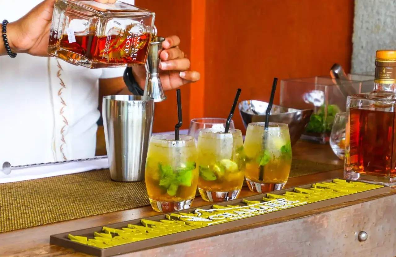 Barman przygotowuje drink z martini, nalewając rum do shakera. Obok stoją gotowe drinki z miętą i lodem.