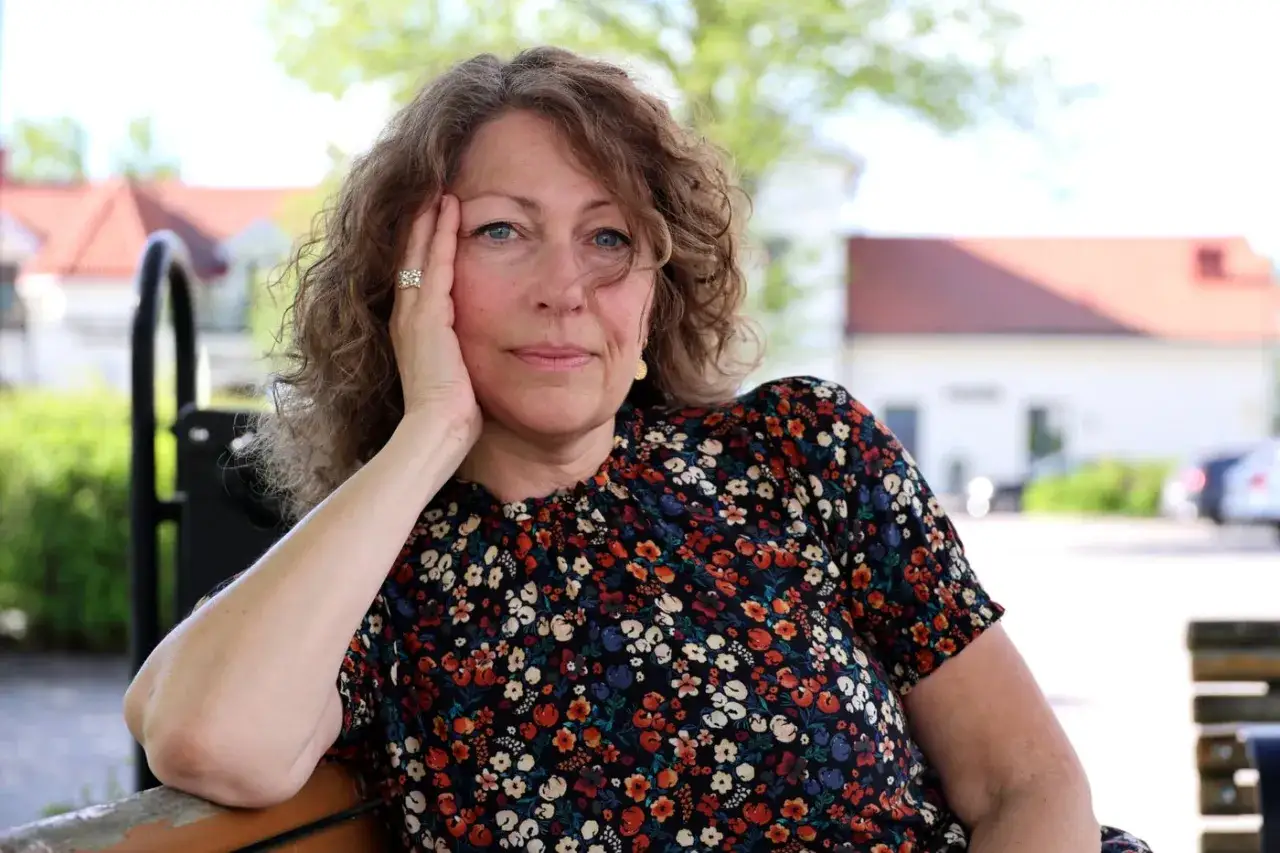 Elisabeth Åsbrink sitter på en bänk med handen mot kinden, klädd i en blommig klänning.