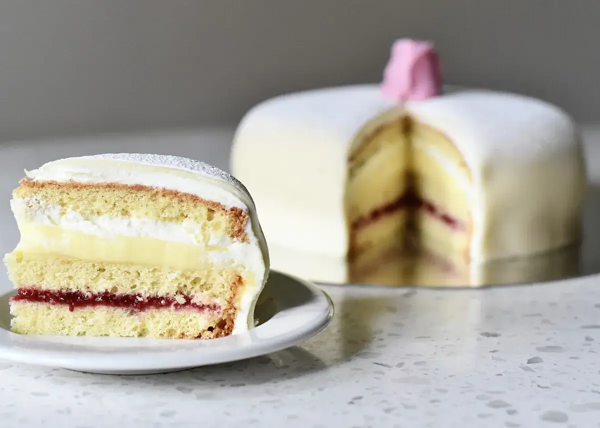En skiva av en klassisk prinsesstårta med sylt, grädde och vaniljkräm. Hela tårtan syns i bakgrunden, dekorerad med en rosa ros.