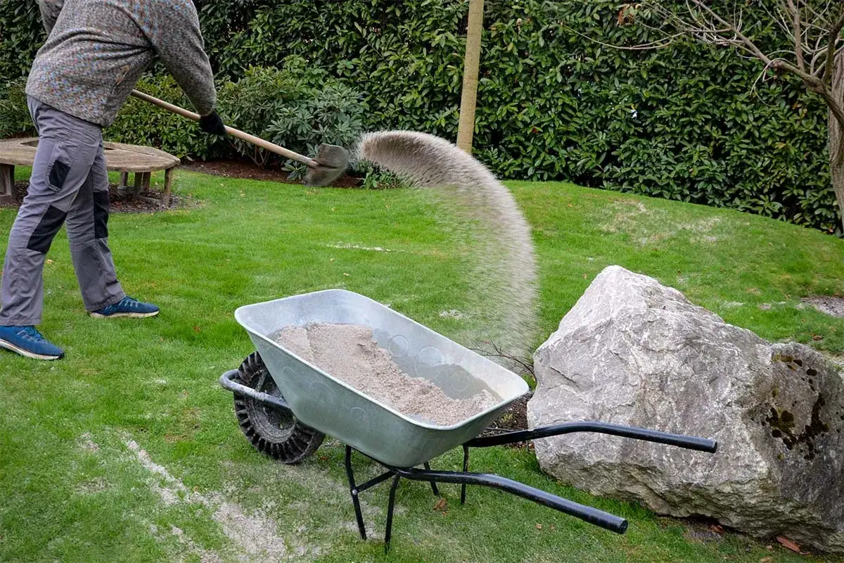 Herbstliche Gartenarbeit: Sand wird mit einer Schaufel aus einer Schubkarre auf den Rasen gesch&uuml;ttet.