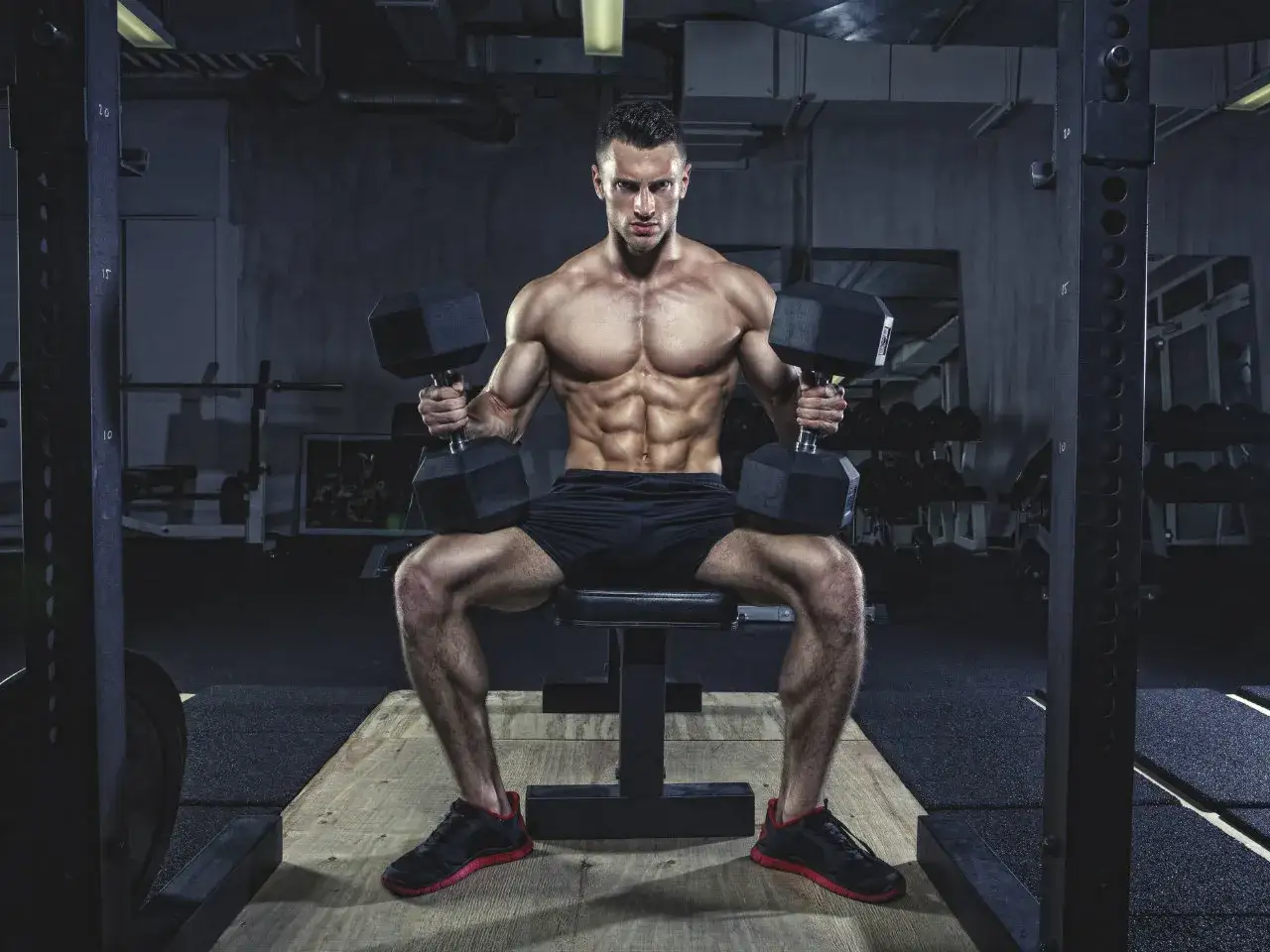 Mężczyzna z wyrzeźbionym brzuchem wykonuje full body workout z hantlami w siłowni.