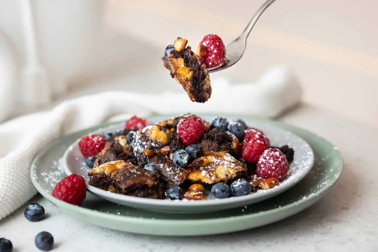 Ein Teller mit Kaiserschmarrn, garniert mit Himbeeren und Blaubeeren. Ein St&uuml;ck wird mit einer Gabel herausgehoben.