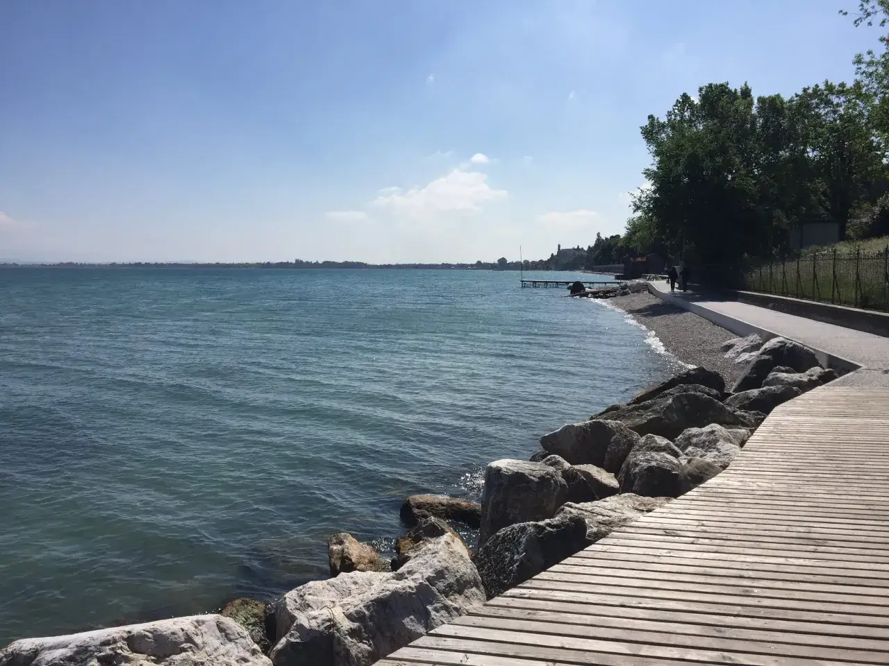 Passeggiata sul lungolago di Desenzano, con acqua azzurra, rocce e un pontile in legno.