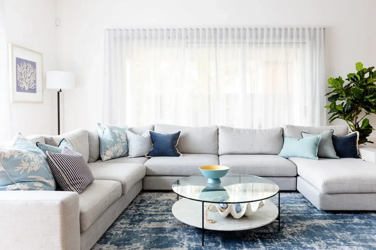 Gem&uuml;tliches Wohnzimmer im Hamptons-Stil mit gro&szlig;em Ecksofa, Kissen in Blau- und Wei&szlig;t&ouml;nen, Glastisch und einem Teppich mit abstraktem Muster.