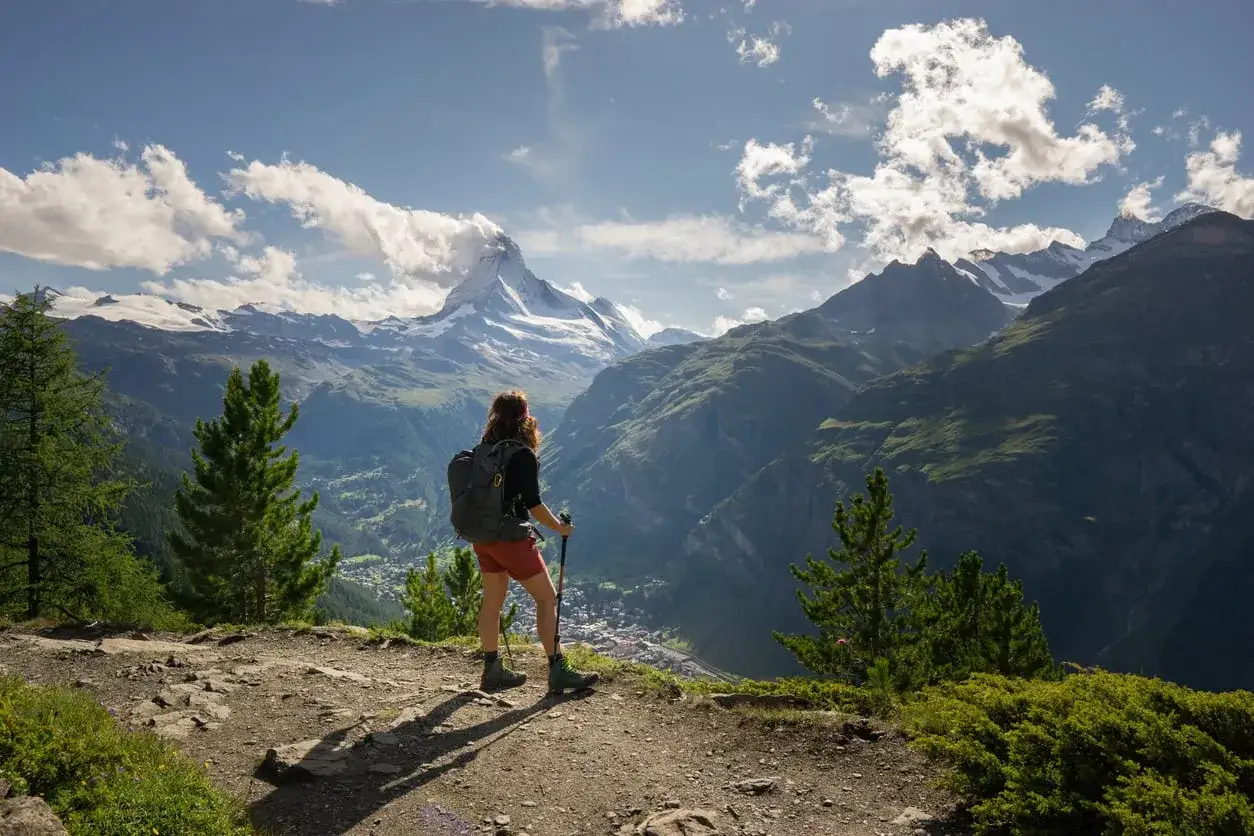 Widok na majestatyczny Matterhorn z perspektywy turystki na szlaku. W oddali widać dolinę i miasteczko, a krajobraz zapiera dech w piersiach, idealny na **Haute Route**.