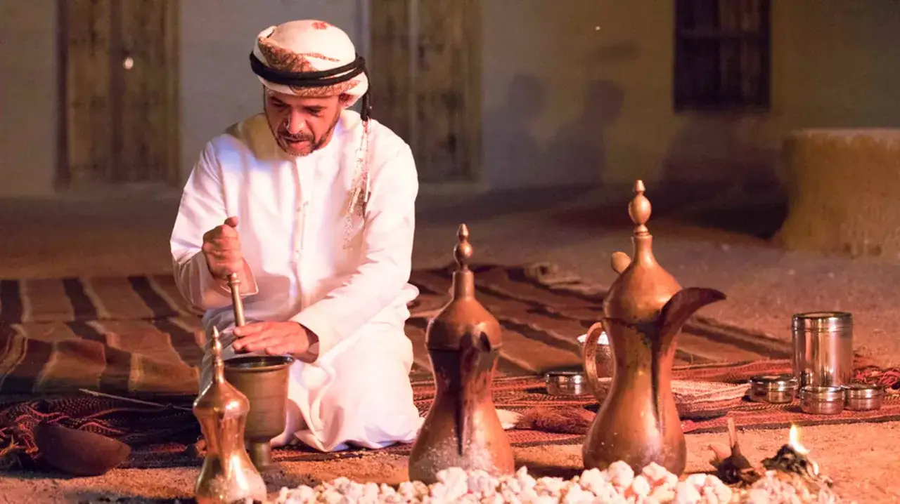 Uomo in abito tradizionale prepara il **caff&egrave; arabo** pestando spezie in un mortaio, circondato da caffettiere e braci ardenti.