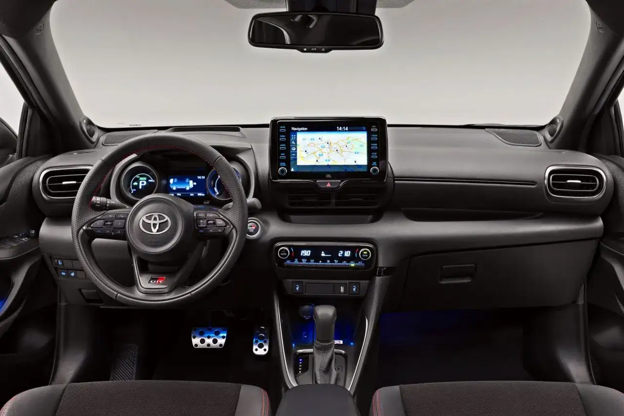 Wnętrze Toyota Yaris GR Sport: kierownica z czerwonymi przeszyciami, ekran nawigacji, cyfrowe zegary i sportowe pedały.