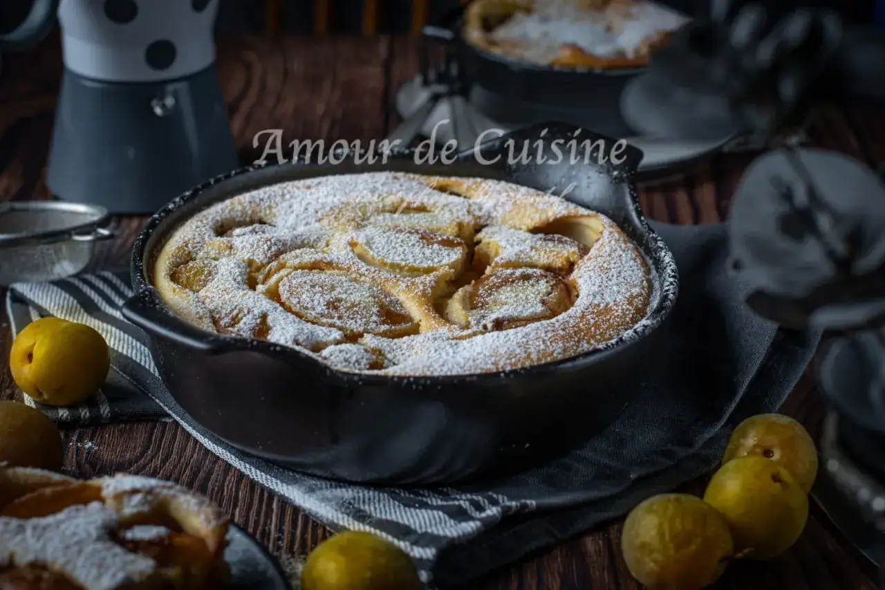 Un d&eacute;licieux clafoutis aux prunes, saupoudr&eacute; de sucre glace, dans un plat en fonte. Quelques prunes fra&icirc;ches l'accompagnent.