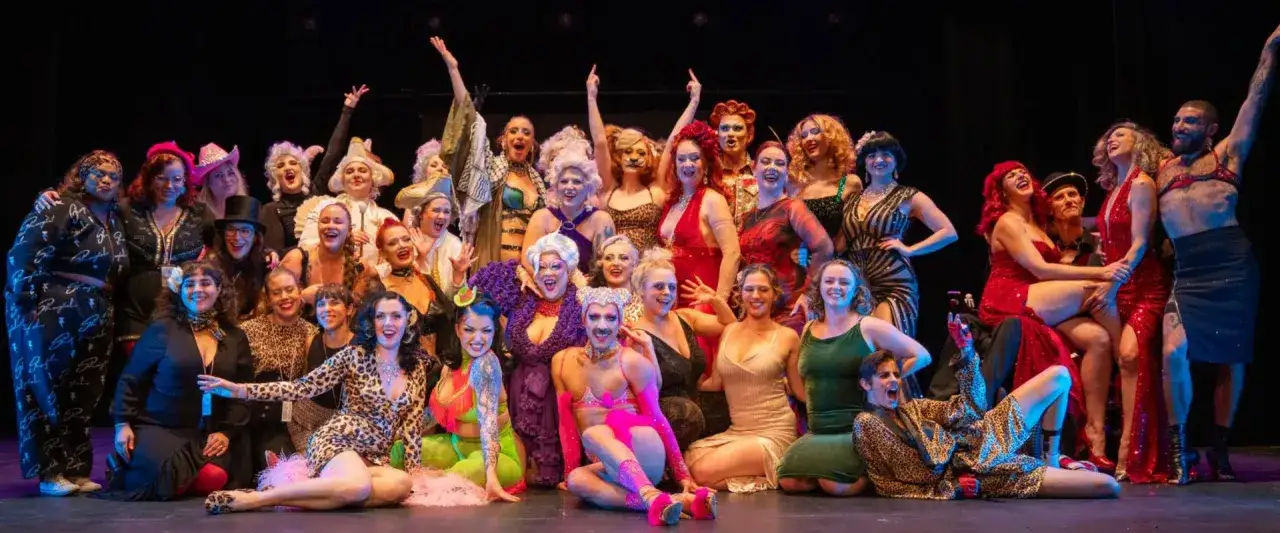 En stor grupp glada artister i fantasifulla kostymer poserar på scenen under Stockholm Burlesque Festival.