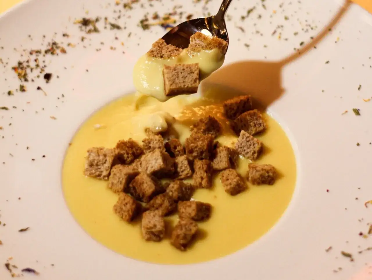 Un cucchiaio solleva cubetti di pane croccante da una cremosa fonduta Valle d'Aosta, guarnita con erbe aromatiche.