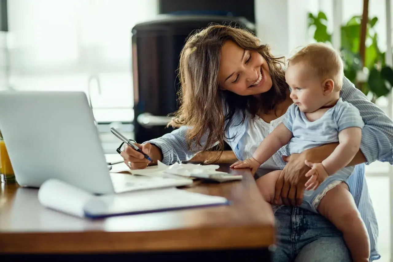 Uśmiechnięta mama z dzieckiem na rękach pracuje przy laptopie. Koszty utrzymania dziecka, nawet 12000 na dziecko, to wyzwanie.
