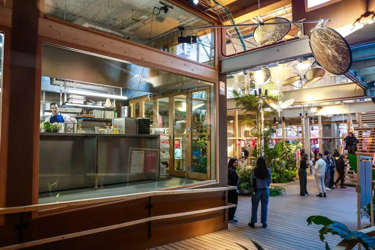 Un chef prepara alimentos en un moderno negocio de comida. Gente observa el ambiente, con plantas y luces decorativas.