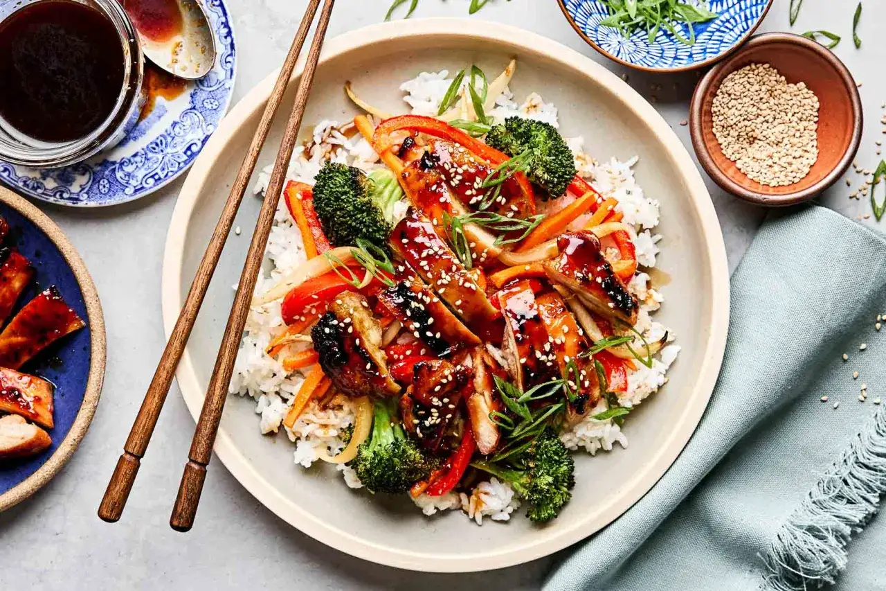 Ein Teller mit Reis, belegt mit saftigem Hähnchen Teriyaki, knackigem Brokkoli, Paprika und Zwiebeln, garniert mit Sesam und Frühlingszwiebeln.