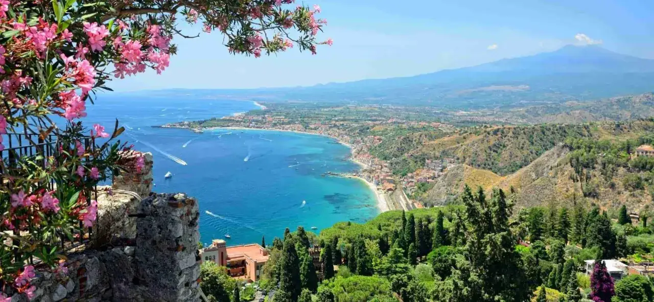 Vista panoramica di Giardini Naxos Taormina con il mare azzurro, la costa e l'Etna sullo sfondo. Fiori rosa in primo piano.