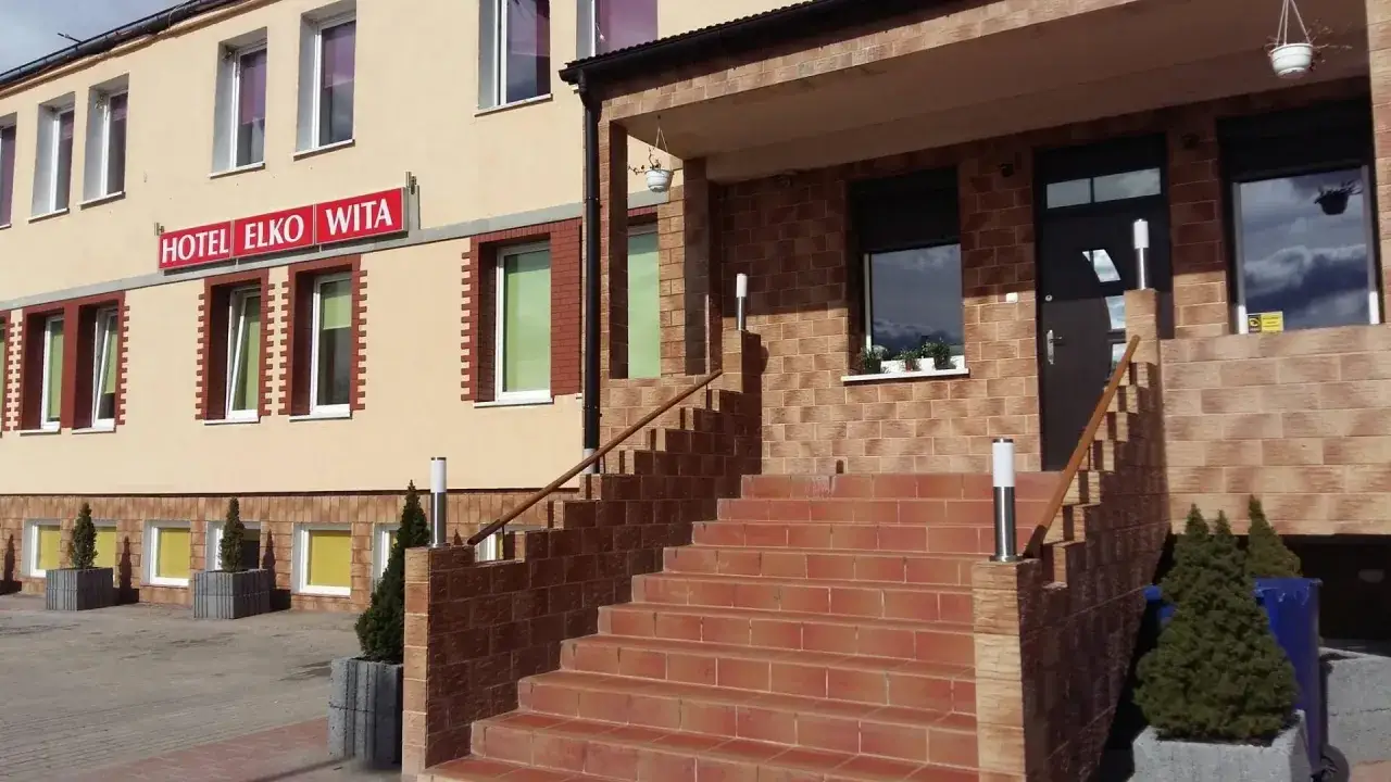 Wejście do hotelu Elko Elbląg. Schody prowadzą do drzwi, a nad nimi widnieje czerwony neon z nazwą obiektu.