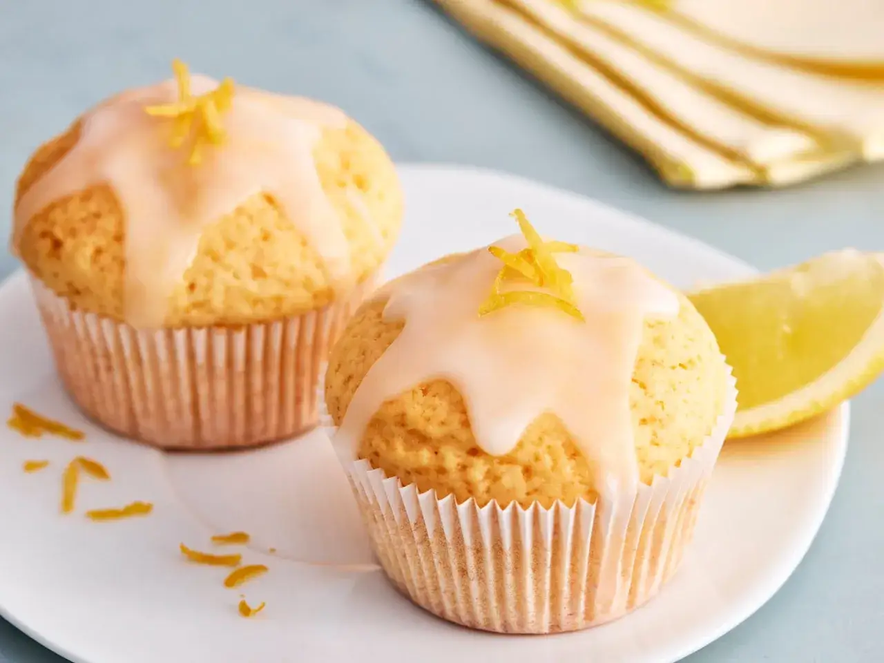 Zwei einfache Zitronenmuffins mit Zuckerguss und Zitronenschale garniert, daneben ein Zitronenschnitz.