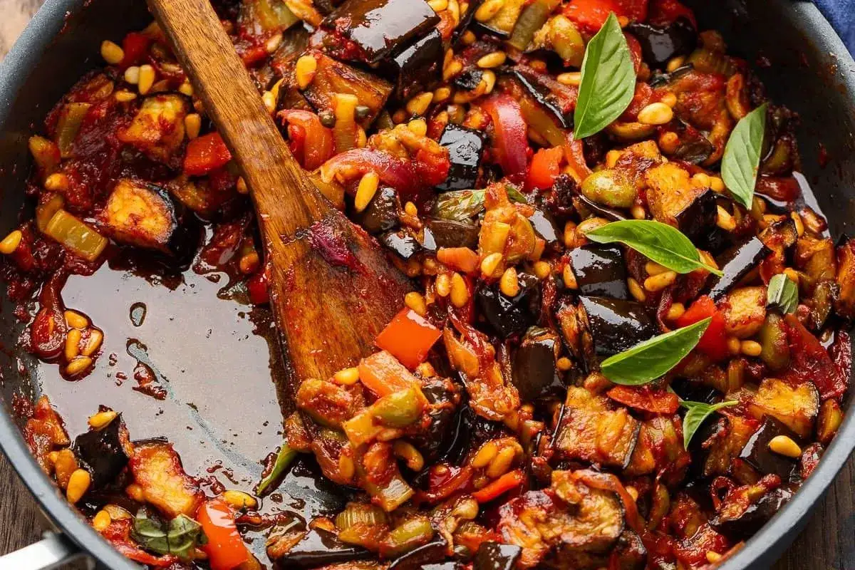 En n&auml;rbild av en panna med en mustig caponata, full av aubergine, paprika och pinjen&ouml;tter. En tr&auml;sked r&ouml;r om i den l&auml;ckra blandningen.