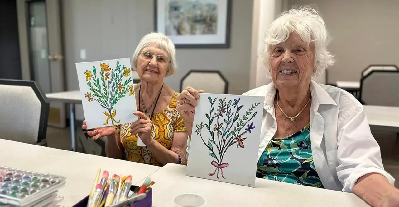 Due anziane sorridono mostrando i loro dipinti floreali, un esempio di attività per anziani con demenza che stimola la creatività.