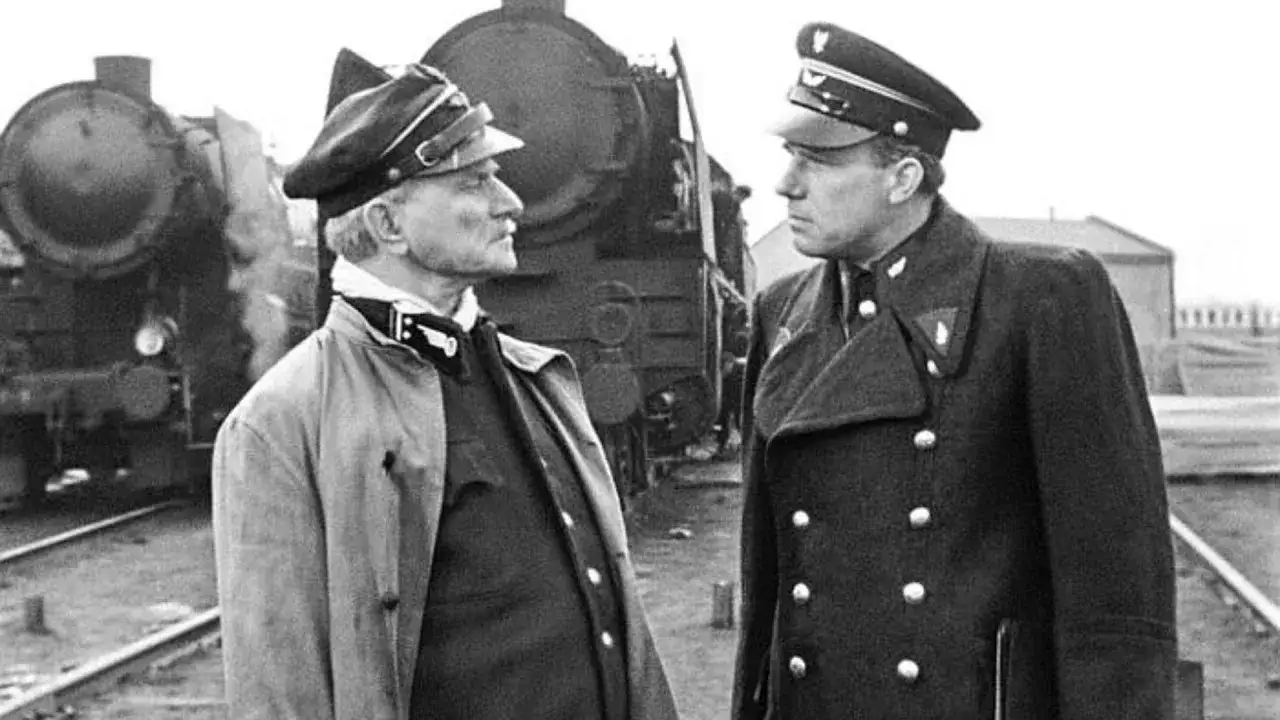 Dwóch mężczyzn w mundurach stoi na tle parowozu. Jeden z nich, starszy, ma na sobie jasny płaszcz i charakterystyczny kołnierz. Drugi, młodszy, w ciemnym płaszczu, patrzy na niego z uwagą. To scena z filmu 
