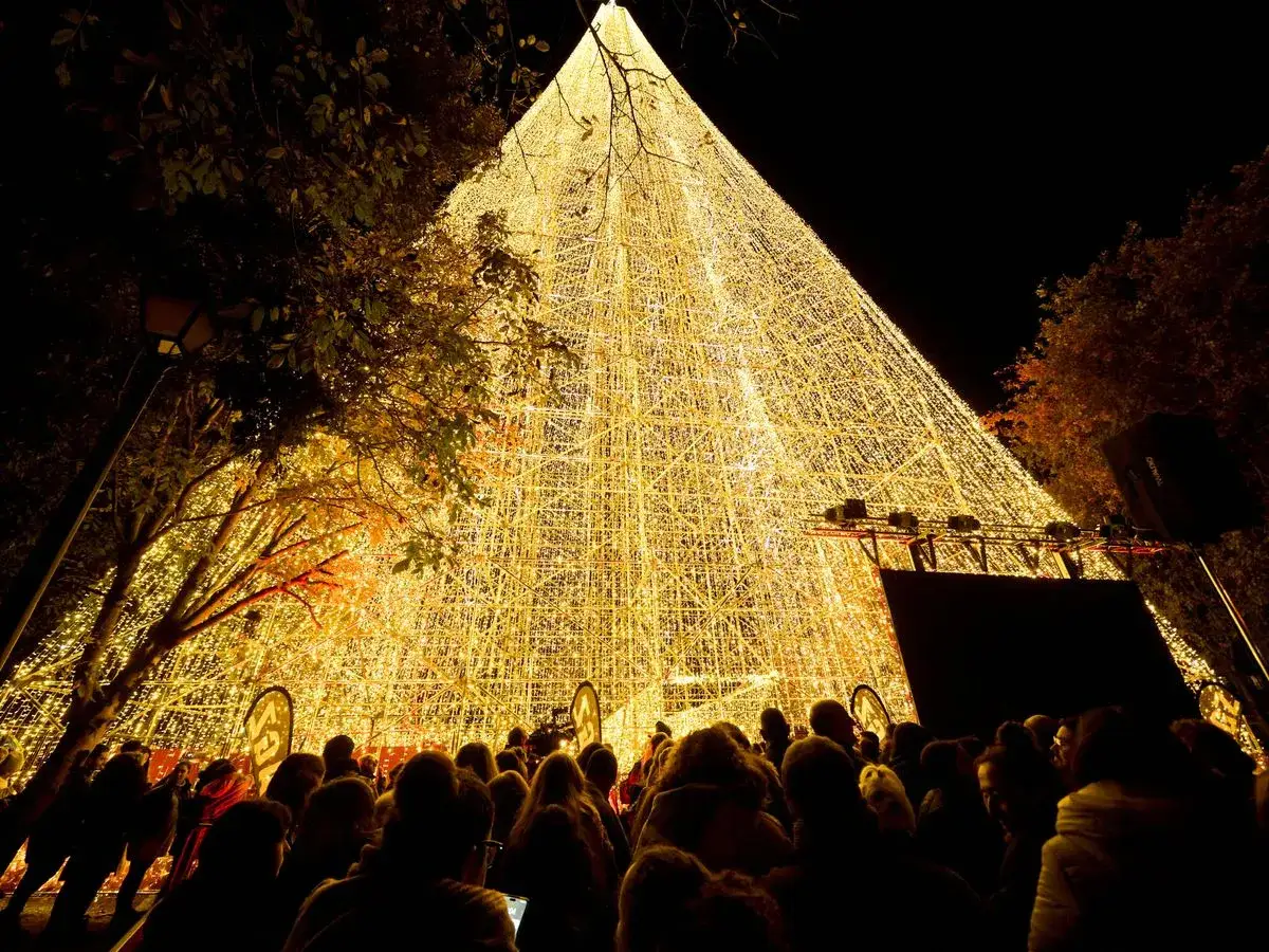 Multitud admira un árbol gigante de luces doradas en un mercado navideño.