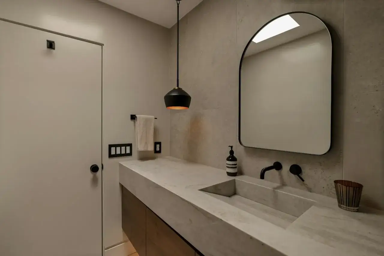 Salle de bain moderne avec lavabo en béton ciré, miroir arqué, porte blanche et luminaire suspendu.