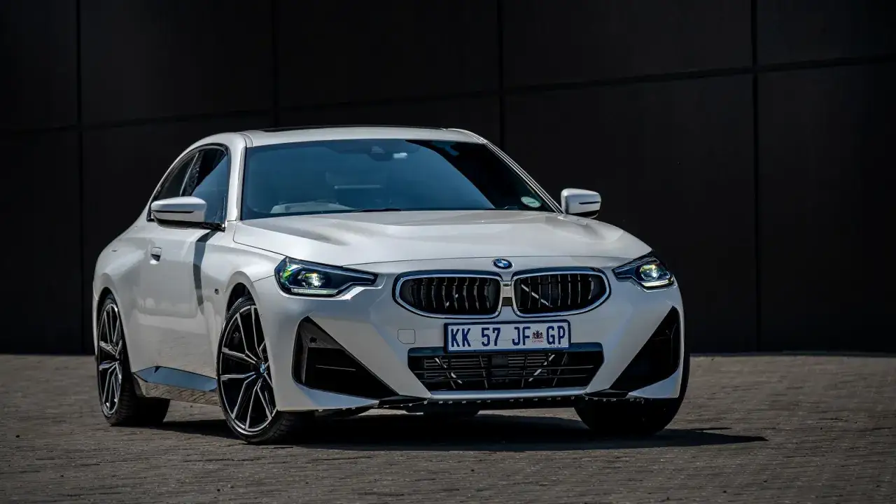 Białe BMW 220i z czarnymi felgami stoi przed ciemną ścianą.