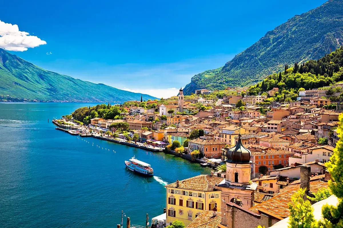 Vista panoramica di Limone sul Garda, con case colorate, il lago blu e montagne verdi. Una nave solca le acque.