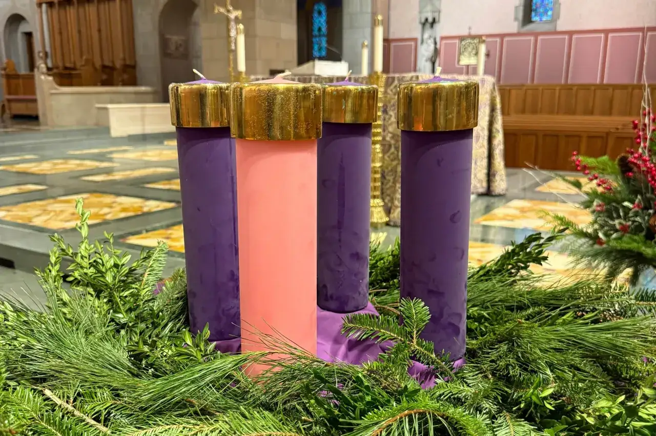 Four Advent candles, three purple and one pink, sit in a wreath of evergreen branches, ready for the 4th Advent.