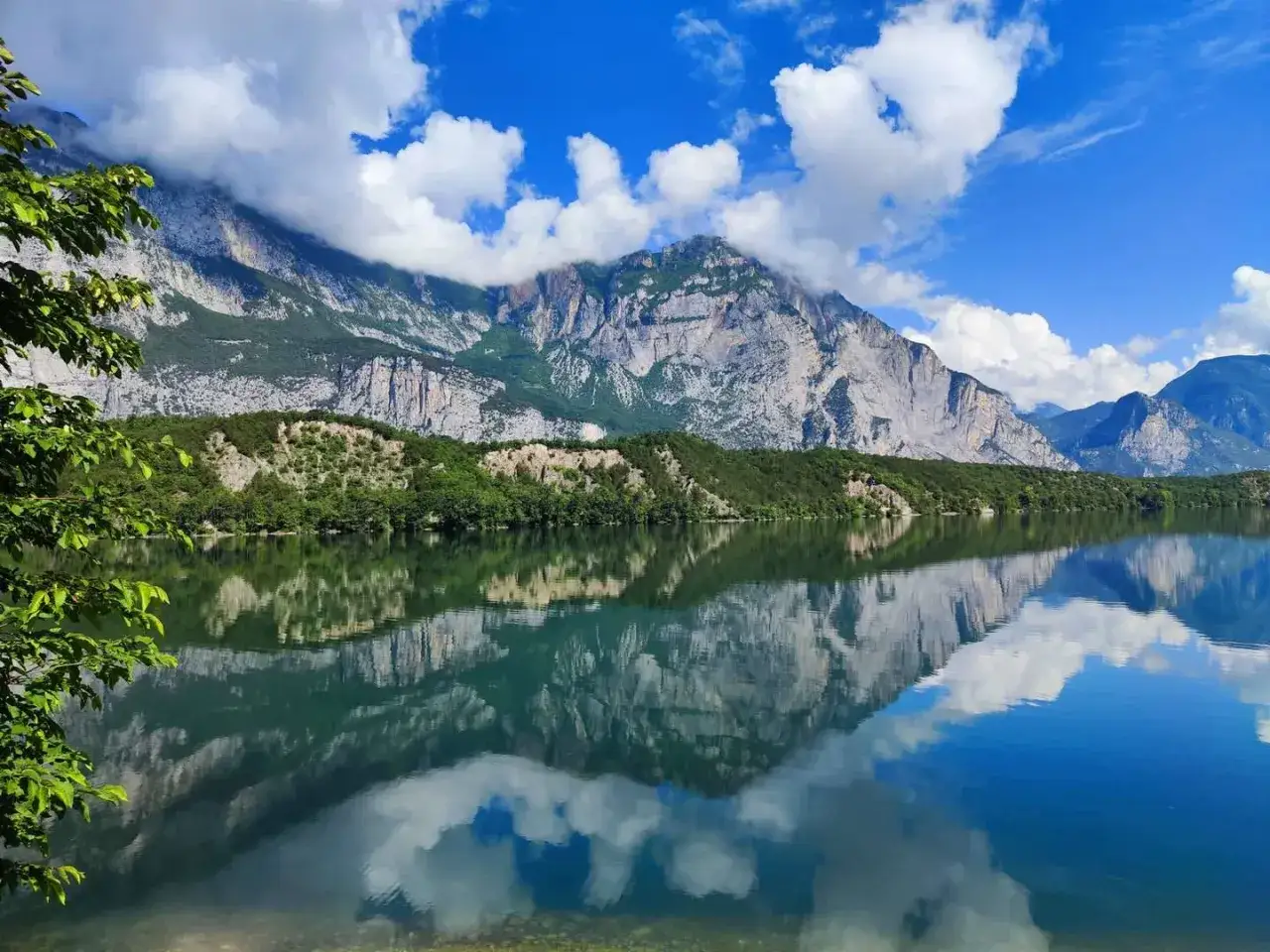 Il **lago di Cavedine** riflette montagne imponenti e nuvole bianche in un cielo azzurro.